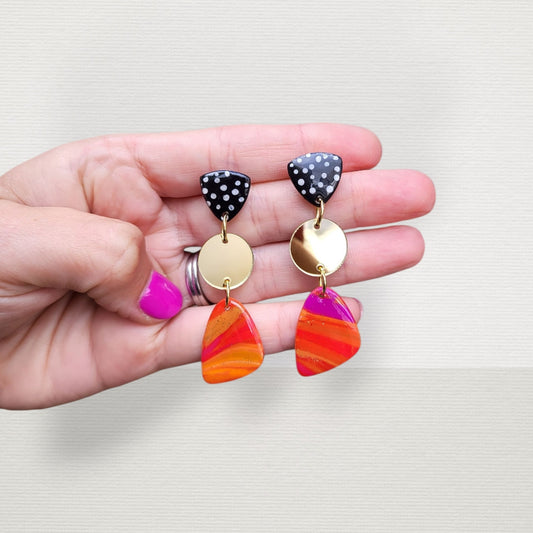 Pink, orange and gold dangling earrings with black and white polka dot details.
The design begins with a sleek, polished polka-dot stud, leading down to a playfully textured fuchsia pink piece. Below, a glossy green charm, accented with luxurious gold leaf, adds a touch of elegance. The earrings finish with a cascade of multi-colored, textured teardrops and gold-flecked 